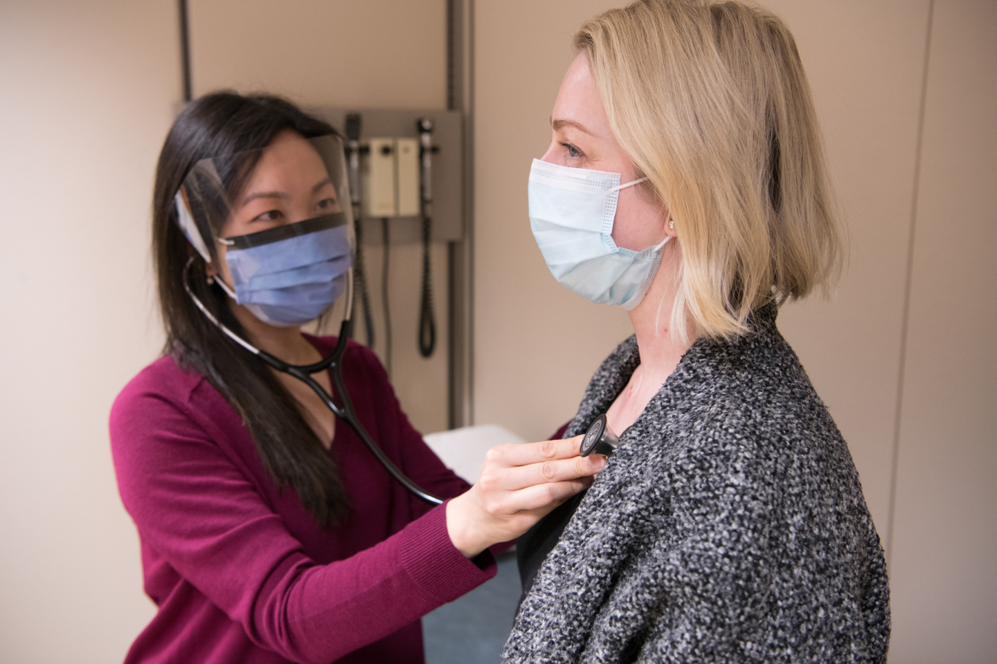 A doctor wearing a mask examines a masked patient with a statoscope